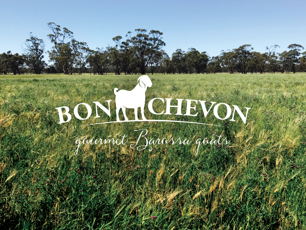 A lush green field with tall grass and scattered trees under a clear blue sky. The white silhouette of a goat and the words "Bon Chevon Gourmet Barossa Goats" are superimposed in the center of the image.