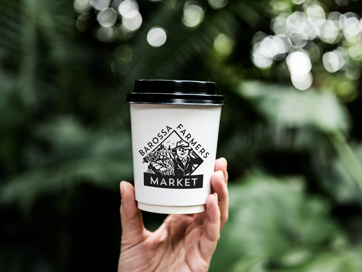 A hand holding a white paper coffee cup with a black lid. The cup has "Barossa Farmers Market" printed on it, along with an illustration of vegetables. The background is blurred greenery.