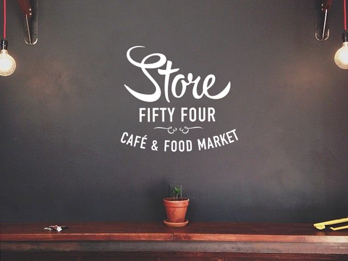 A minimalist café interior features a dark wall with the text "Store Fifty Four Café & Food Market" in a stylish font. Above a wooden counter sits a small potted plant, flanked by two hanging light bulbs, creating a cozy ambiance.